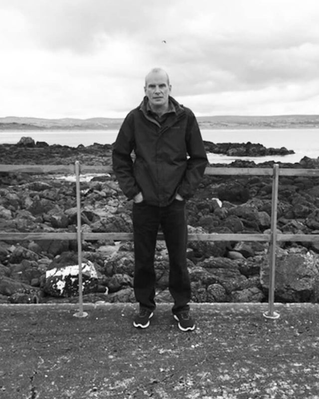 a black and white photo of a man standing in front of a railing at the seaside. He looks happy.